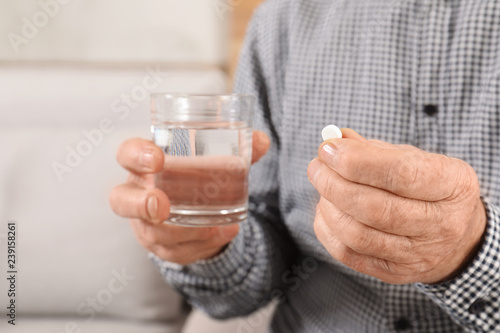 Senior man holding pill and glass of water indoors, closeup