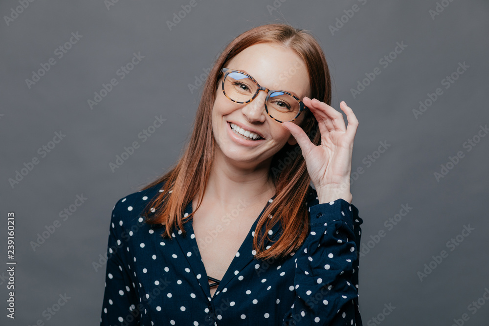 Smiling woman with pleased facial expression, keeps hand on rim of spectacles, wears black polka dot blouse, rejoices promotion at work, poses over grey background. People, happiness concept Smiling woman with pleased facial expression, keeps hand on rim of spectacles, wears black polka dot blouse, rejoices promotion at work, poses over grey background. People, happiness concept