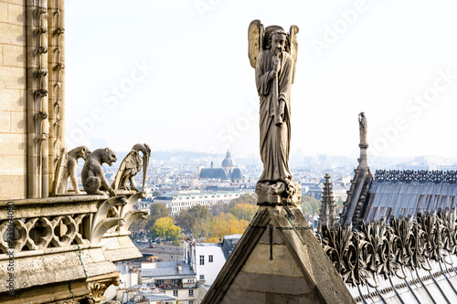Stone statue of an angel with trumpet on the rooftop of Notre-Dame de Paris cathedral and three chimeras overlooking the city, with church of Saint-Paul-Saint-Louis, vanishing in the mist.
