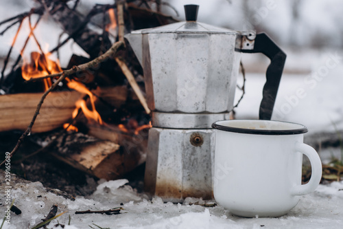  Сup of coffee and a coffee maker on the background Camp fire an in winter time, surrounded by snow against the near of the frozen lake. Concept adventure active vacations outdoor hiking sport