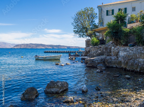 Pebble sea shore and The White House in Kalami, former residence of British authors Lawrence and Gerald Durrell and their family. Boat and pier at stone quay. Summer blue sky, corfu Greece
