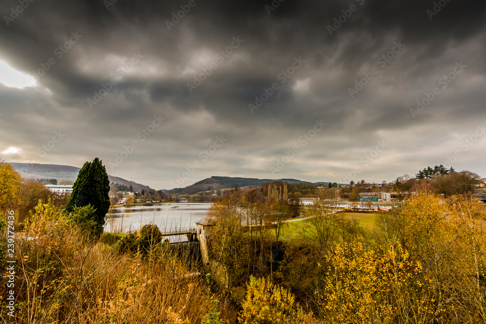 Landscape Doyards lake with Vielsalm village and mountains in background against sky with stormy clouds, view from a hill among autumn trees, cloudy day in the Belgian Ardennes