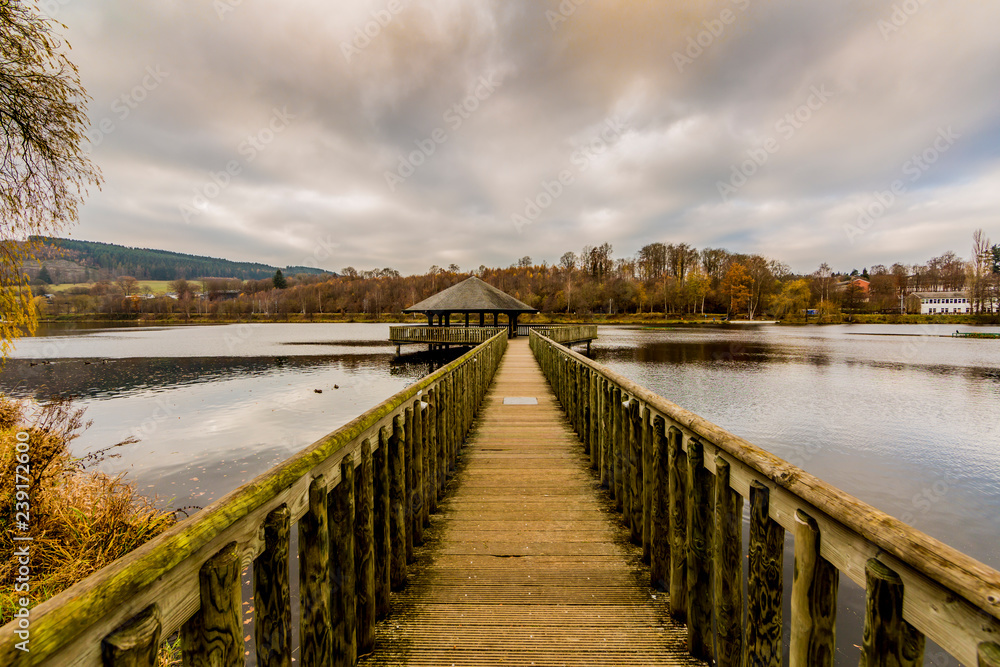 Straight wooden bridge towards a gazebo in the middle of Lake Doyards surrounded by autumn greenery, light wind on water surface, cloudy sky covered with clouds at Vielsalm in the Belgian Ardennes