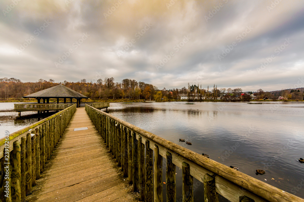 Wooden bridge leading to a gazebo in the middle of the Doyards lake surrounded by autumn vegetation, light wind on the water surface, cloudy sky covered with clouds in Vielsalm, the Belgian Ardennes