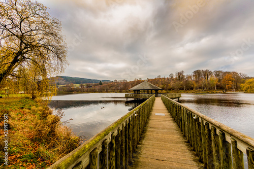 Straight wooden bridge leading to a gazebo in the middle of the Doyards lake surrounded by autumn vegetation, cloudy sky covered with clouds in Vielsalm, the Belgian Ardennes