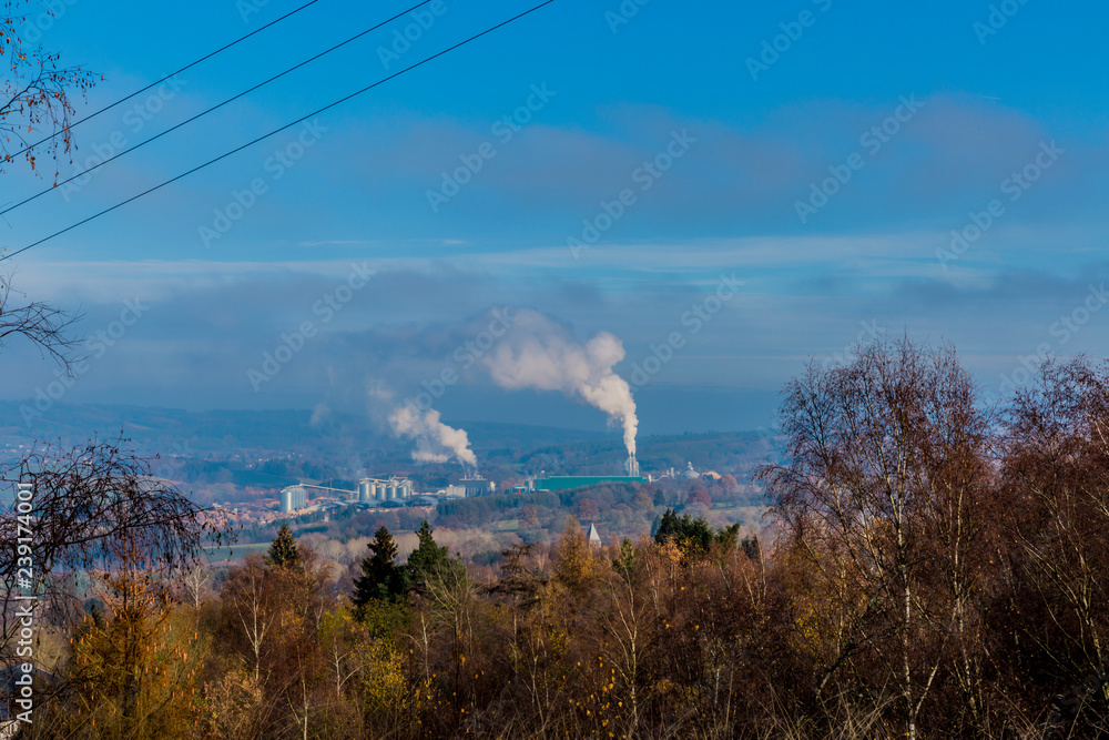 Industrial complex emitting smoke or steam against blue sky, seen from a hill with abundant brown trees in forest of the Belgian Ardennes, sunny winter day in Belgium. Environmental pollution concept