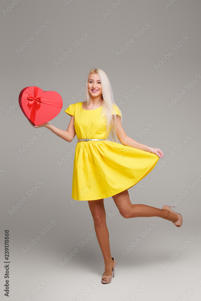 Blond girl in yellow dress with heart shape box for Valentine Day Holiday on grey background. Copy space for text Blond girl in yellow dress with heart shape box for Valentine Day Holiday on grey background. Copy space for text
