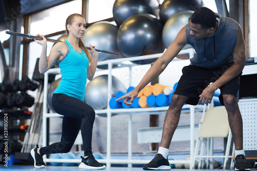 African-american trainer consulting young active woman during exercise with gymnastic bar in gym