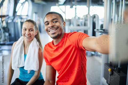 Happy young intercultural man and woman making selfie in fitness center at break after training