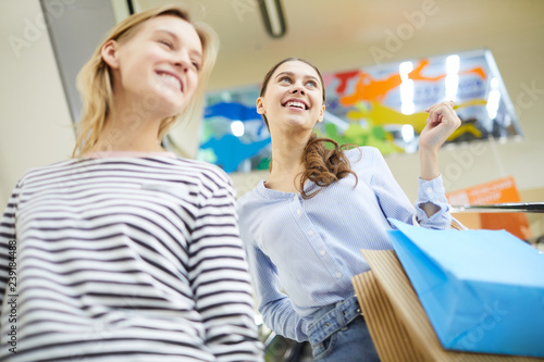 Two young cheerful customers with bags enjoying time in large modern mall at seasonal sale