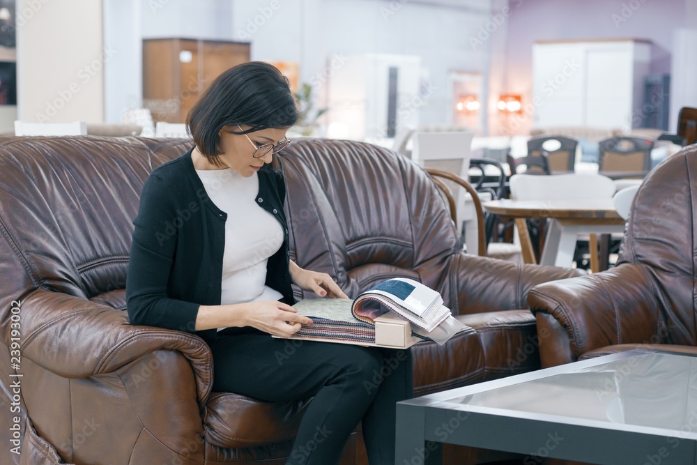 Adult woman buyer looking at a book with upholstery fabrics, female is sitting on brown leather sofa in furniture showroom shop Adult woman buyer looking at a book with upholstery fabrics, female is sitting on brown leather sofa in furniture showroom shop