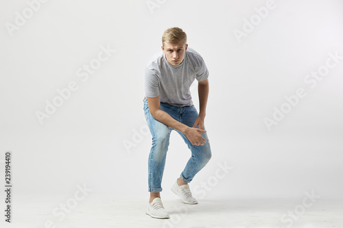 Serious blond guy dressed in a white t-shirt and jeans stands with his hands on the knees on the white background in the studio © Leika production