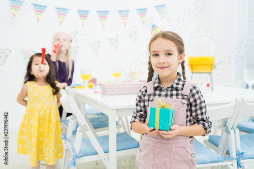 Smiling content pretty girl in party hat standing against table and holding blue gift box while looking at camera in decorated room photo