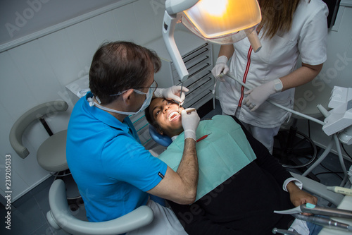Dentist examining oral cavity of young African-American man working in in dental clinic with assistant from above.