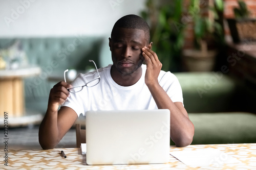 Tired African American man taking off glasses, feel eye strain after long work with laptop, computer, exhausted overworked student in cafe, sitting with closed eyes having bad sight vision problem