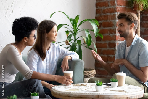 Diverse friends meeting in coffee house talking at table, friendly man telling story to women, gesticulate, workmates or colleagues spending time together in cafeteria during break, enjoying coffee