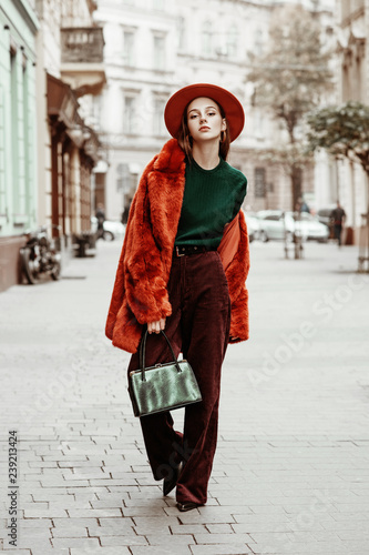 Outdoor full body fashion portrait of young beautiful woman wearing trendy oversized orange faux fur coat, hat, green sweater, corduroy trousers, holding stylish snakeskin bag, posing in street 