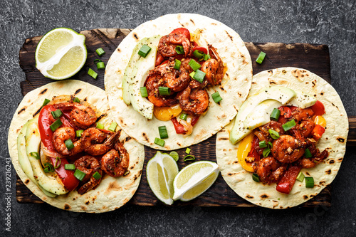 Fajitas in tortillas with fried shrimps, bell peppers and onion served up with avocado and green onions on wooden cutting board, top view, flat lay