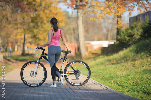 Young beautiful girl in sportswear stands next to a bicycle on the street in the park in autumn.