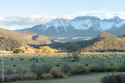 The sun begins going down over partly cloud hidden snow capped mountain ranges above autumn colored farm land and forests photo