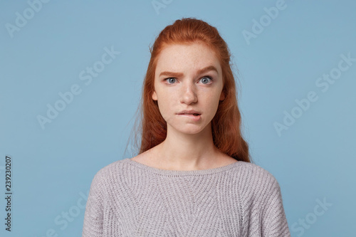 Young beautiful red-haired teen girl looks concerned puzzled in a panic, one eyebrow raised bit her lower lip isolated on a blue background. photo