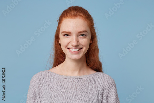 Headshot of attractive cheerful red-haired young woman smiling pleasantly and looking at the camera isolated against a blue background. photo