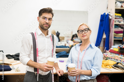 Waist up portrait of two mature tailors looking at camera while posing in atelier holding coffee cups