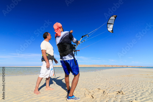 Two men on sea shore sand beach learning to pilot training kite in blue sky with wind, recreation leisure vacation fun active hobby. Sunny summer warm day. Kite boarding water sports lessons course