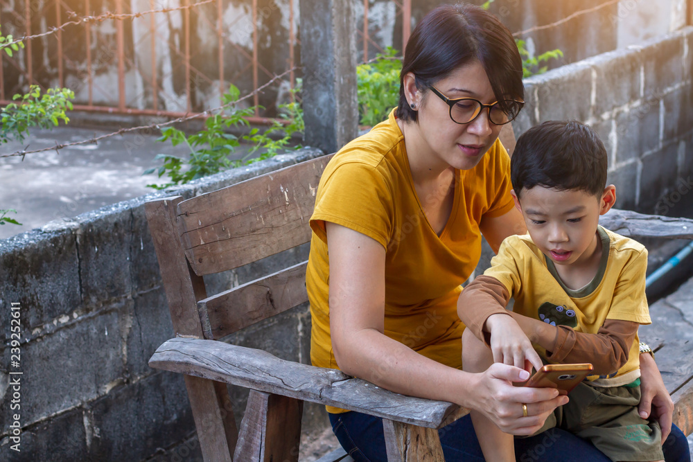 Asian black hair boy age 4 years old interested content in his mother’s smart phone with sunset effect. Mom teaching son using mobile phone background. Selective focus and copy space Asian black hair boy age 4 years old interested content in his mother’s smart phone with sunset effect. Mom teaching son using mobile phone background. Selective focus and copy space