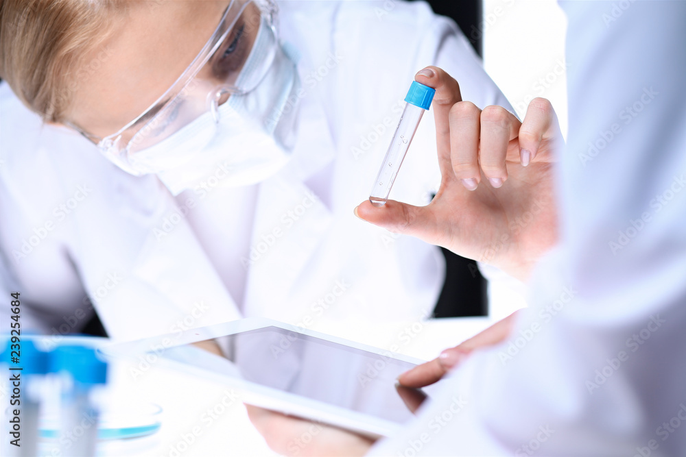 Closeup of scientific research team with clear solution in laboratory. Blonde female chemist holds test tube of glass while her colleague checks results with tablet pc. Blood test, medicine or Closeup of scientific research team with clear solution in laboratory. Blonde female chemist holds test tube of glass while her colleague checks results with tablet pc. Blood test, medicine or