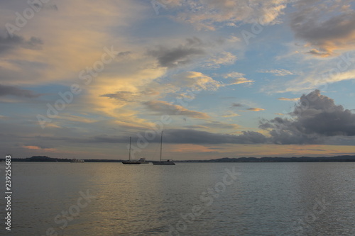 Calm surface of Waitemata Harbour reflecting sun light with moored sailing boats and distant land in background in cloudy evening. © Daniel Poloha