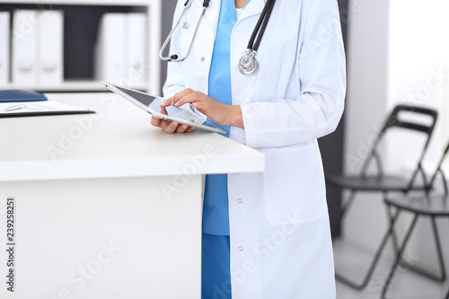 Close-up of female physician using digital tablet while standing near reception desk at clinic or emergency hospital. Unknown doctor woman at work. Medicine and healthcare concept