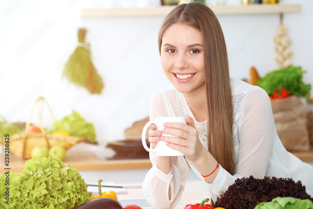 Young happy woman holding white cup and looking at the camera while sitting at wooden table in the kitchen among green vegetables. Good morning, lifestyle or cooking concept