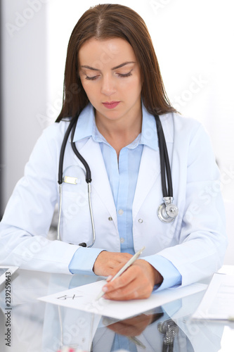 Doctor woman at work. Portrait of female physician filling up medical form while sitting at the glass desk at clinic or hospital. Medicine and healthcare concept