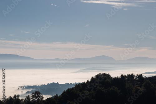 Fog filling a valley in Umbria (Italy), with layers of mountains and hills, trees in the foreground and various shades of blue
