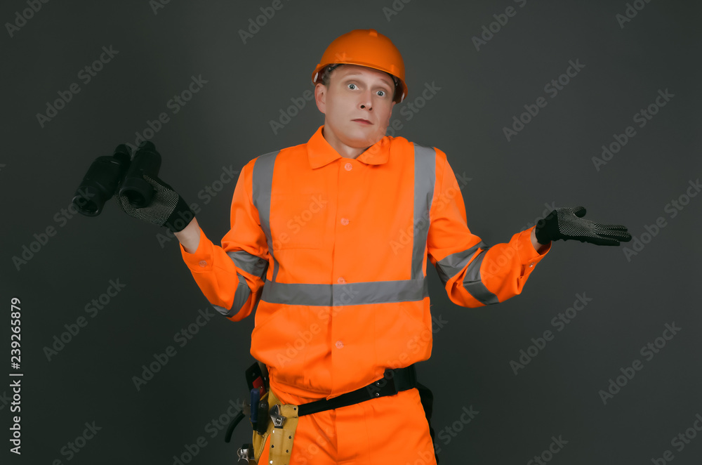 Geodesist or builder foreman with a binoculars is shrugging by his shoulders isolated on gray background. Geodesist or builder foreman with a binoculars is shrugging by his shoulders isolated on gray background.