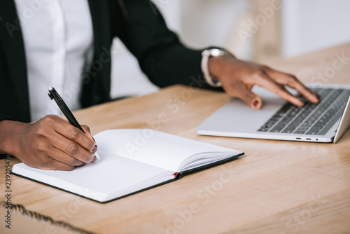 cropped view of african american woman writing in notebook and using laptop