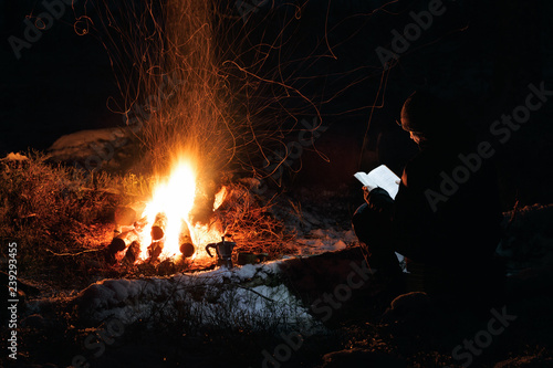 Man reading a book by the fire. Book is highlighted by headlamp Winter night in the forest