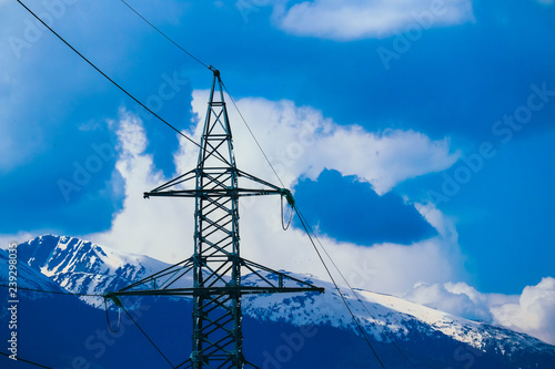 High-voltage powerful electricity tower with wires, alpine ice mountains peaks  on background.