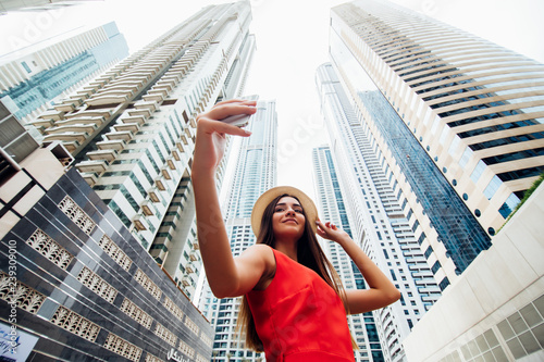Photography Young woman in red dress and summer hat take selfie photo on the phone with modern skycrapers on background