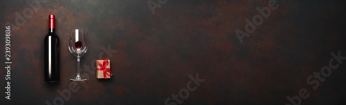 Bottle of wine with wineglass and gift box on rusty brown background. Panoramic top view with space for your text