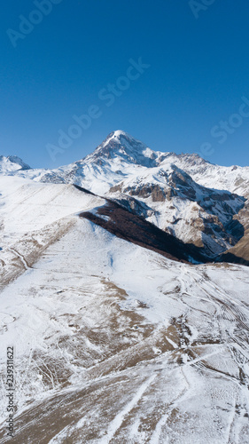 Aerial photo of caucasus winter mountainous landscape with mountains ranges. It is winter snowy mountains in Kazbegi, Georgia. There are snow dunes, hills and clear blue sky. Georgian nature.