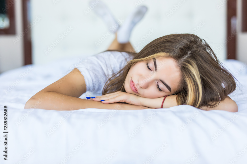 Young woman lying at the end of the bed looking forward while smiling and her head resting on both of her hands. Young woman lying at the end of the bed looking forward while smiling and her head resting on both of her hands.