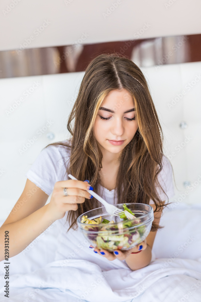 Young woman lying on bed eating vegetable salad