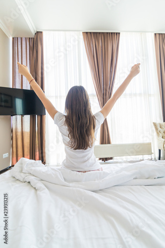 Young woman outstretching her arms sitting on the bed after good night sleep, unwilling to get up and leave her comfortable nest, entering a day happy and relaxed, ready for productive work. Rear view