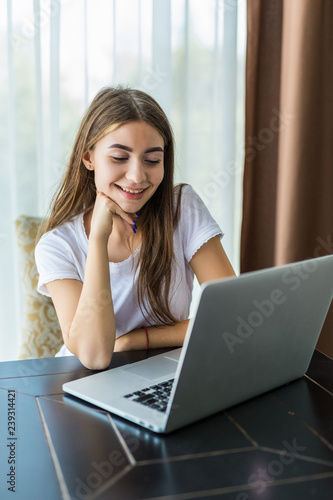 Young smiling girl is sitting on modern chair near the window in light cozy room at home working on laptop in relaxing atmosphere