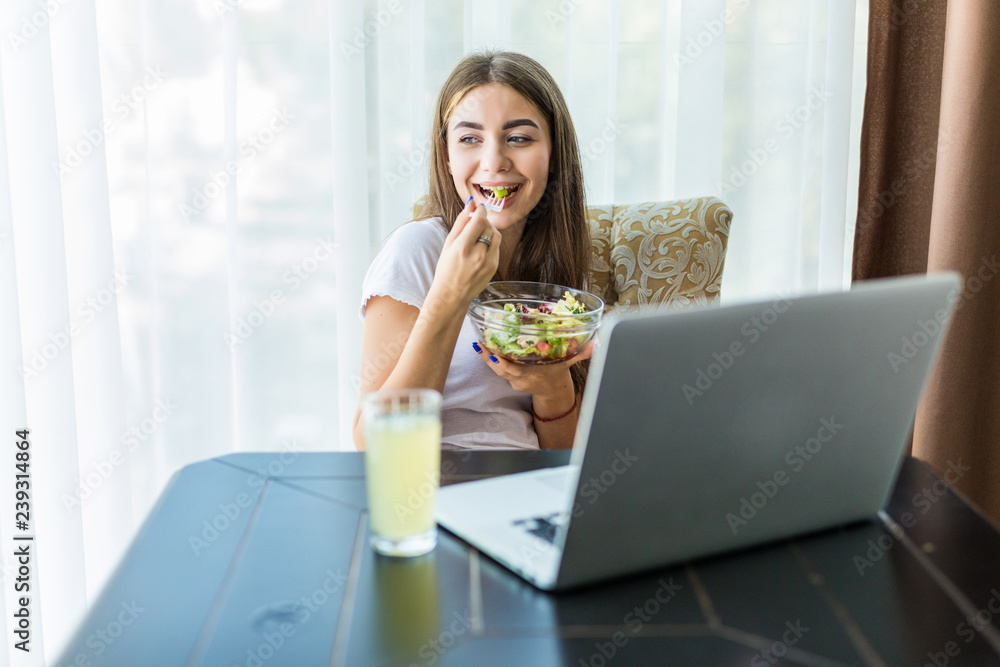 Young woman with salad and orange working on laptop sitting at desk at home Young woman with salad and orange working on laptop sitting at desk at home