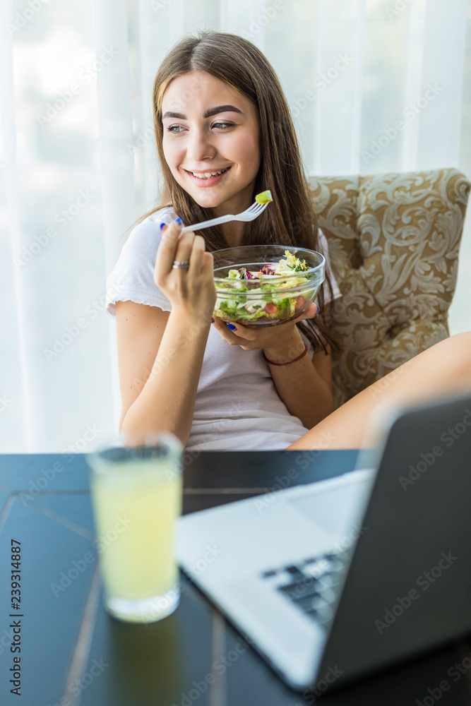 Portrait of a happy young woman eating fresh salad from a bowl while sitting and using laptop computer at home Portrait of a happy young woman eating fresh salad from a bowl while sitting and using laptop computer at home