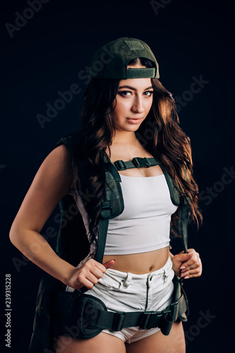 Porter sexy brunette woman with long hair in short white shorts and a top, military elements and a baseball cap on a black background © liliyabatyrova