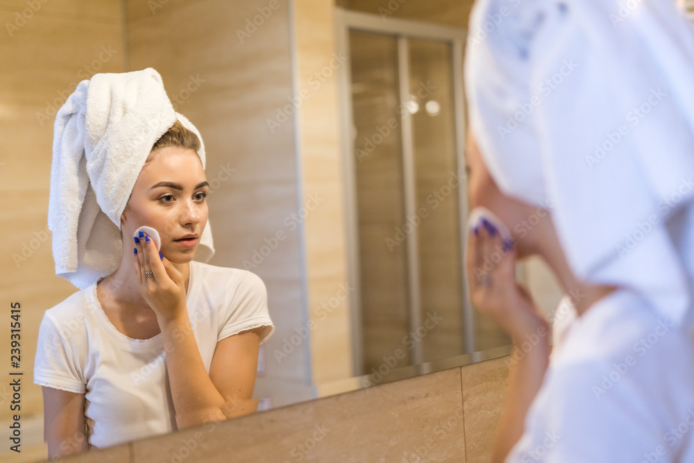 Fototapeta premium Beautiful young woman is treating her face after shower standing and touching her cheek with a sponge. The woman is holding a mirror and smiling